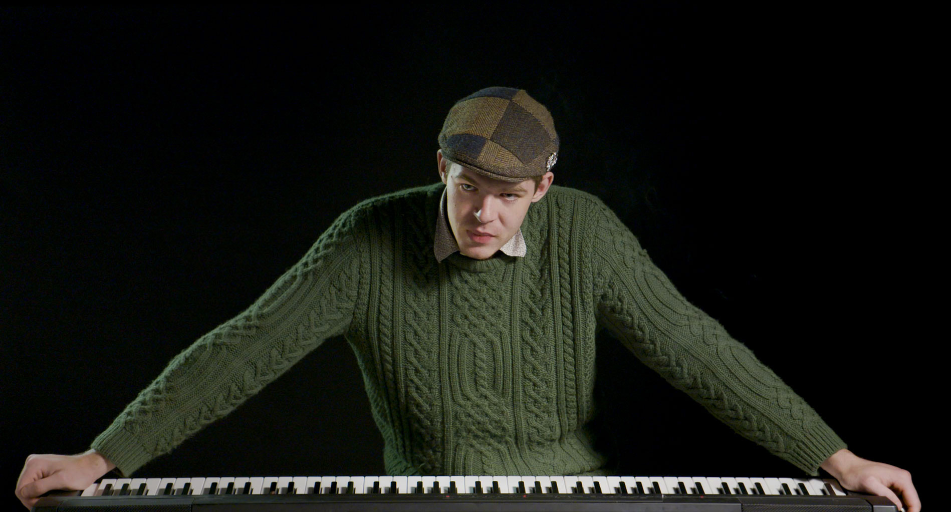 Sean, standing arms wide over a piano keyboard, wearing a flat cap, looking at the camera.